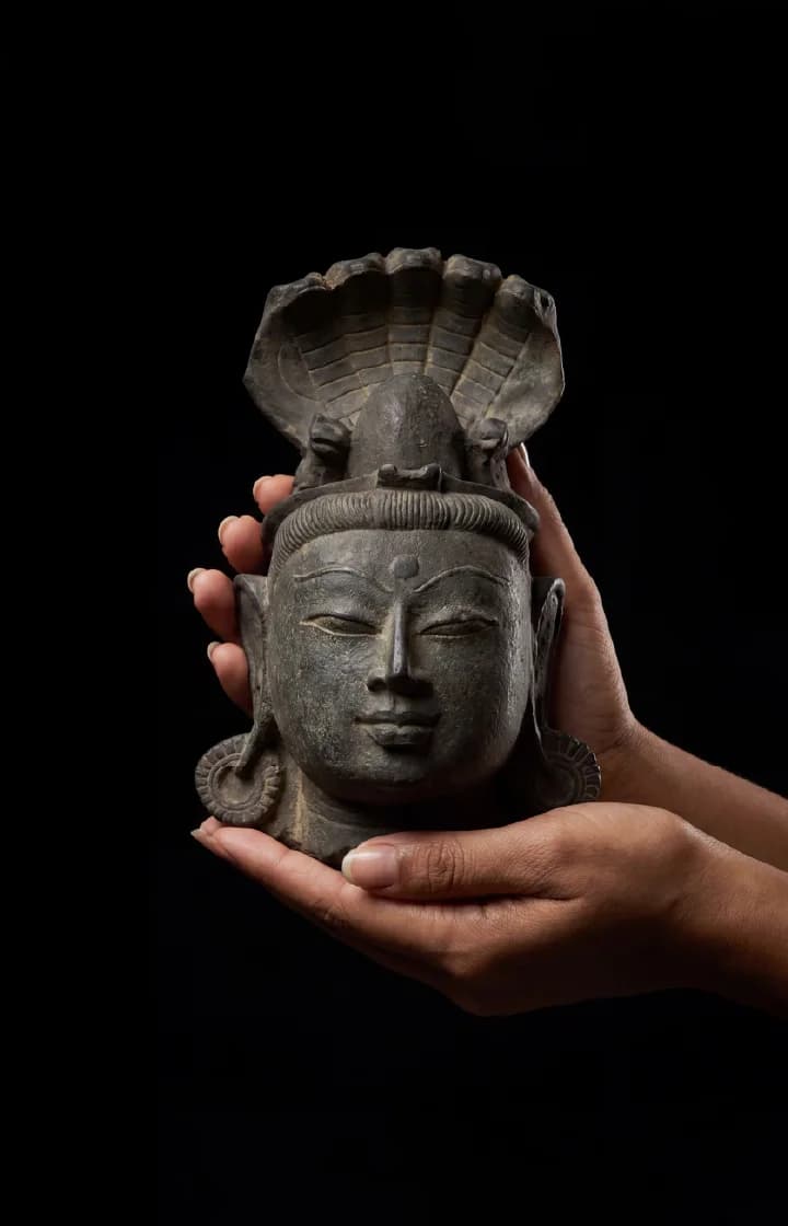A pair of hands gently cradles a dark stone Shiva Lingam with Nandi & Serpent replica against a black background.