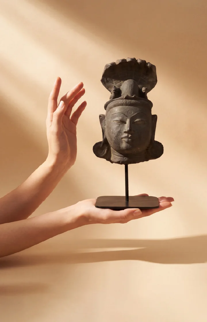A pair of hands gently cradles a dark stone Shiva Lingam with Nandi & Serpent replica on a minimalist stand.