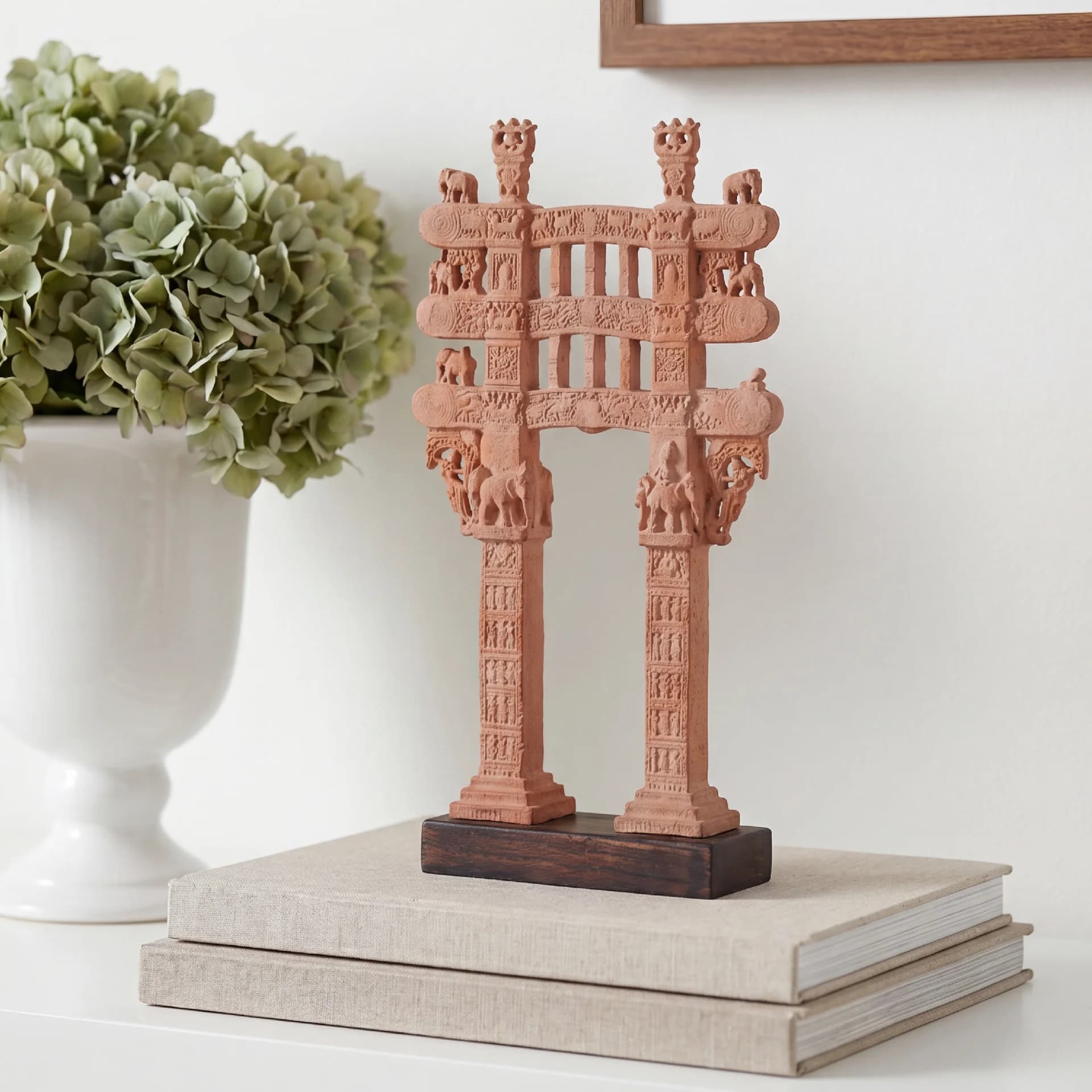A terracotta Sanchi Torana Gateway Miniature displayed on stacked books next to a white vase with green flowers.